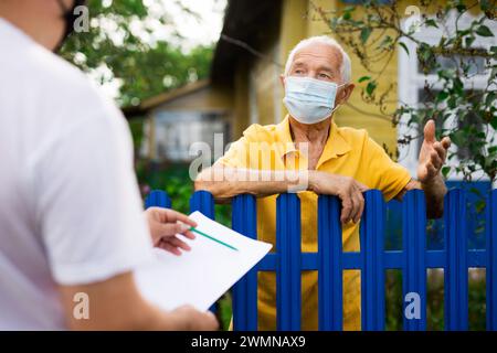 Älterer Mann in Schutzmaske, der mit dem Vertreter der Versicherungsgesellschaft kommuniziert, während er am Zaun seines Landhauses steht Stockfoto