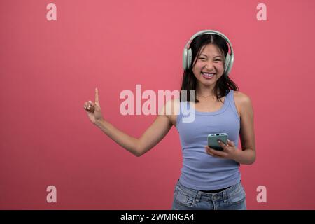 Fröhliche Frau, die Radio hört und das Telefon nach oben zeigt. Junge glückliche Frau, die Kopfhörer verwendet, hören Musik mit Smartphone isoliert auf rotem Hintergrund Stockfoto
