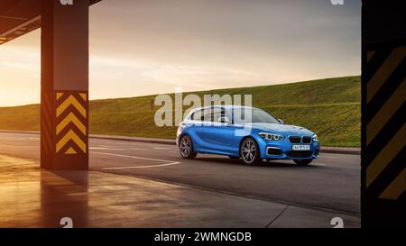 Blauer BMW 1er (F21) auf einem Parkplatz bei Sonnenuntergang. Dreiviertelansicht der sportlichen BMW M140i Fließheck mit zwei Säulen im Vordergrund Stockfoto