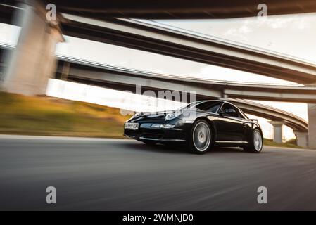 Bewegungsaufnahme des schwarzen Porsche 911 Carrera Coupés. Porsche 997 fährt bei Sonnenuntergang unter der Brücke. Stockfoto