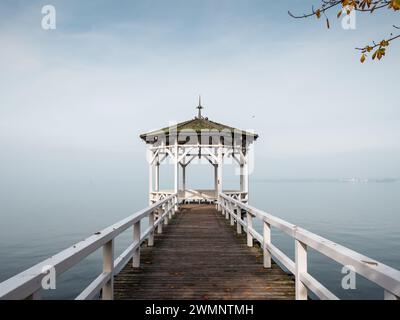 Herbstszene auf der Seepromenade: Symmetrisches Foto des weißen Pavillons am Ufer des Sees, bedeckt mit Nebel. Stockfoto