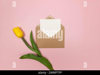 Draufsicht auf gelbe Tulpe, Kraftumschlag und weiße Karte auf rosa Hintergrund. Farbenfrohe Komposition im Frühling, flacher Lay, Kopierraum. Stockfoto