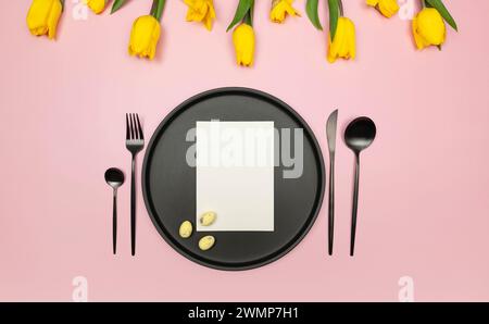 Blick von oben auf gelbe Tulpen, Narzissen, ostereier auf rosa Hintergrund. Tischaufstellung, schwarzer Teller und Besteck, weiße Karte. Frühjahrs-osterkomposition. Stockfoto