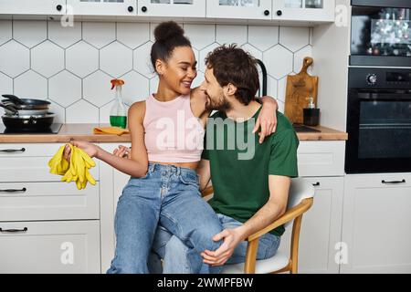 Liebevolles, fröhliches, multirassisches Paar, das auf einem Stuhl sitzt und sich mit Lappen und Handschuhen in den Händen umarmt Stockfoto
