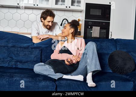 Fröhliche attraktive afroamerikanische Frau, die auf dem Sofa sitzt und ihren fröhlichen Freund ansieht Stockfoto