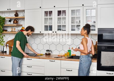 Ansprechendes, fröhliches interrassisches Paar in Hauswäsche, das Hausaufgaben macht und nach dem Frühstück die Küche putzt Stockfoto