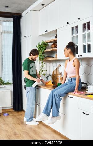 Eine fröhliche afroamerikanische Frau, die auf der Theke sitzt und ihren Freund bei der Arbeit beobachtet Stockfoto