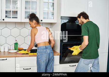 Fröhlich schönes interrassisches Paar in Haushaltswaren, das ihre Küchenutensilien aufmerksam zu Hause reinigt Stockfoto