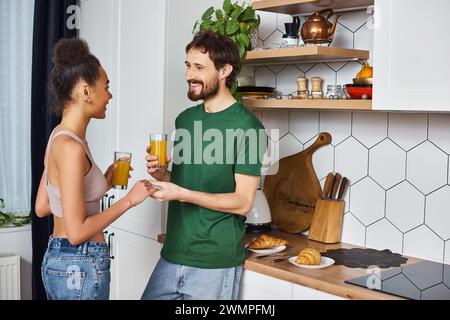 Ein ansprechendes, abwechslungsreiches Paar in Haushaltswaren, das Orangensaft trinkt und sich anlächelt Stockfoto