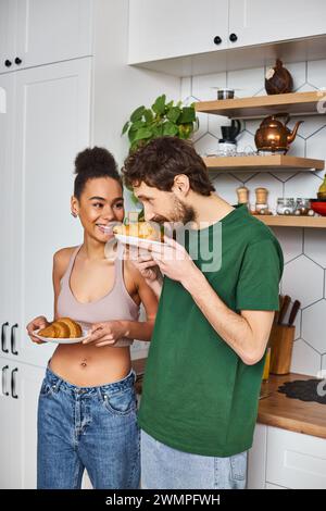 Gut aussehend fröhliches, abwechslungsreiches Paar in gemütlichen Haushaltswaren, das Teller mit Croissants in der Küche hält Stockfoto