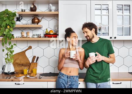 Ansprechendes, fröhliches, abwechslungsreiches Paar in Homewear, das heissen Kaffee genießt und sich ansieht Stockfoto