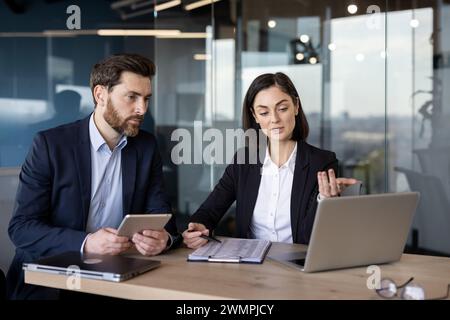Erfolgreiche Geschäftspartner sitzen mit digitalen Geräten am Tisch und schauen aufmerksam auf den pc-Bildschirm. Kaukasische Frau und Mann in Formalkleidung diskutieren und analysieren Statistiken der Verkaufssaison. Stockfoto
