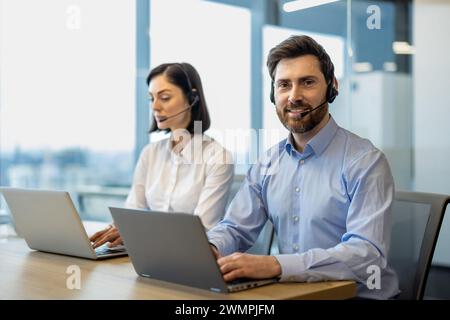 Ein paar Kundenbetreuer, die in Headsets mit Mikrofonen sitzen, die während des Arbeitstages am Laptop sitzen. Bärtiger Mann lächelt in die Kamera, während eine Frau mit kurzem Haarschnitt auf der pc-Tastatur am Schreibtisch schreibt. Stockfoto