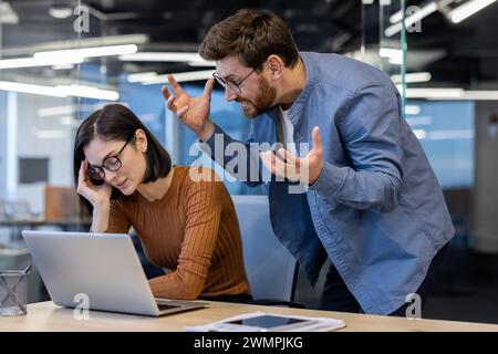 Aggressiver männlicher Boss in Denim-Hemd spreizt die Hände und schreit die genervte Mitarbeiterin am Schreibtisch an. Gestresste Frau, die im Büro vor dem Laptop sitzt und Kritik vom Regisseur nimmt. Stockfoto
