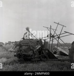 1960er Jahre, historische Erntezeit, draußen auf einem Feld, ein Landwirt, der auf einer Heuballenpresse sitzt und von einem Traktor gezogen wird, der die Griffe der Ballenpresse betätigt, um kleine Strohballen zu produzieren, England, Großbritannien. Stockfoto