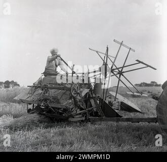 1960er Jahre, historische Erntezeit, draußen auf einem Feld, ein Landwirt, der auf einer Heuballenpresse sitzt und von einem Traktor gezogen wird, der die Griffe der Ballenpresse betätigt, um kleine Strohballen zu produzieren, England, Großbritannien. Stockfoto