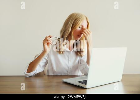 Müde überarbeitete Frau, die an Kopfschmerzen leidet, sich die trockenen Augen reibt und mit einem Laptop arbeitet, der im Büro am Schreibtisch sitzt Stockfoto