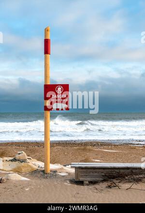 Ein rot-gelbes Schild am Strand warnt Besucher, Eisbären zu beobachten, wenn die Wellen am Ufer der Hudson's Bay in Churchill, Manitoba, Kanada, abstürzen Stockfoto