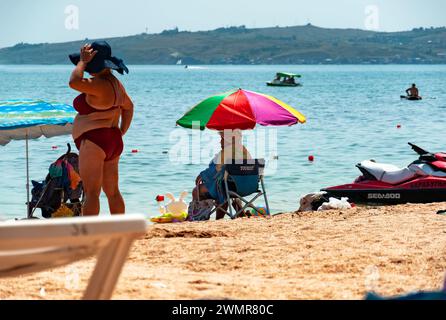 18. August 2021 Republik Krim, Feodosia, Stadtstrand am Schwarzen Meer. Eine ältere Frau sitzt auf einem Stuhl am Meer in der Nähe des Wassers. Stockfoto