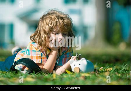 Der Junge spart Geld für den Kauf, hält das rosa Sparschwein, legt sich auf Gras. Kindererziehung zur Berechnung des persönlichen Budgets, Finanzverwaltung und Investitionsförderung Stockfoto