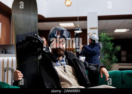 Porträt eines männlichen kaukasischen Touristen, der in der Hotellobby Schneekleidung trägt und Snowboard hält. Das Bild zeigt einen Mann mit Winterkleidung, der in der Lounge des Skigebietes mit Snowboardausrüstung in der Nähe sitzt. Stockfoto