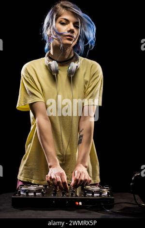 Mädchen, die Kopfhörer tragen, konzentrieren sich auf das Mischen von Musik auf einem Soundboard. Weiblicher DJ mit blauen Haaren und gelbem T-Shirt. Stockfoto