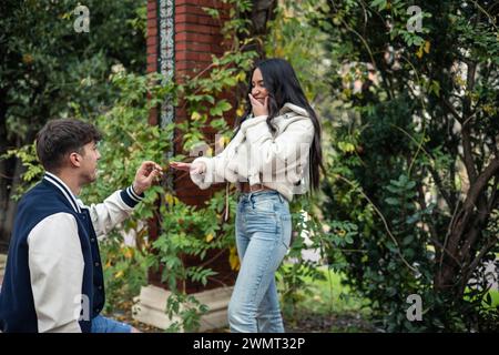 Junger kaukasischer Mann kniet vor seiner überraschten Latina-Freundin in einem Park Stockfoto