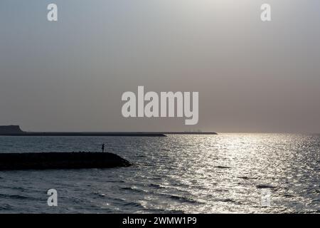 Silhouetten aus Steinbrechern mit einer Person am Ende mit rauem Sonnenlicht, das in den Wellen des Golfs reflektiert, am Strand Al Hamriyah in den Vereinigten Arabischen Emiraten. Stockfoto