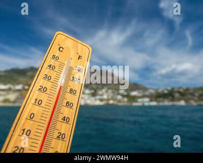 Holzthermometer mit roter Messflüssigkeit, die hohe Temperaturen an sonnigem Tag vor dem Hintergrund des Meeres anzeigt. Konzept von Urlaub, Urlaub, warmes Wetter Stockfoto