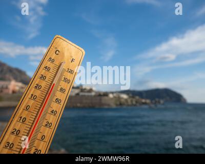 Holzthermometer mit roter Messflüssigkeit, die hohe Temperaturen an sonnigem Tag vor dem Hintergrund des Meeres anzeigt. Konzept von Urlaub, Urlaub, warmes Wetter Stockfoto