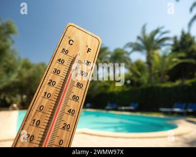 Holzthermometer mit roter Messflüssigkeit, die hohe Temperaturen an sonnigen Tagen auf dem Hintergrund des Schwimmbades anzeigt. Konzept von Urlaub, Urlaub, warm Stockfoto