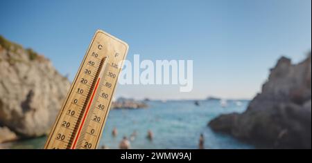 Holzthermometer mit roter Messflüssigkeit, die hohe Temperaturen an sonnigem Tag vor dem Hintergrund des Meeres anzeigt. Konzept von Urlaub, Urlaub, warmes Wetter Stockfoto