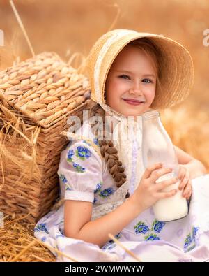 Ein Porträt eines glücklichen Mädchens in einem Weizenfeld bei Sonnenuntergang. Ein Kind hält ein Glas mit Milch vor dem Hintergrund von Roggenohren. Picknick in der Natur. Stockfoto