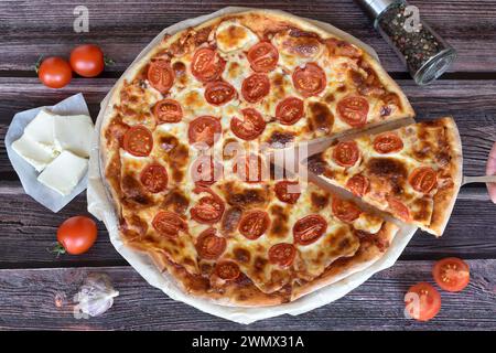 Handschaufel mit Pizzascheibe halten. Frisch gebackene Pizza Margherita auf rustikalem Holztisch. Kirschtomaten, Mozzarella, Pfeffermühle, Knoblauch. Stockfoto