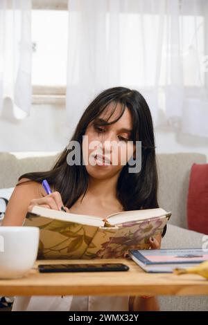 Vertikales Bild der jungen schwarzhaarigen argentinischen Latina Brünette, zu Hause sitzend im Wohnzimmer, lernend, konzentrierend, aufschreibend und einlesend Stockfoto