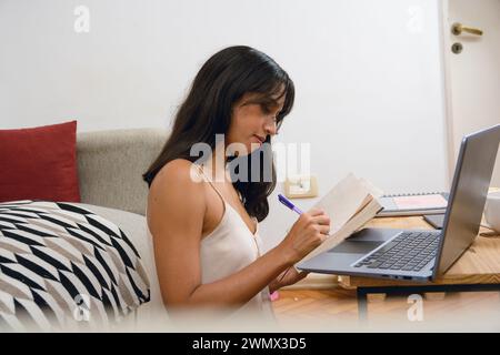 Seitenansicht junge Latina Unternehmerin Brünette Frau mit schwarzen Haaren, glücklich zu Hause sitzend auf dem Boden, beschäftigt sich mit dem Schreiben in ihrem Notizbuch, während sie mit Arbeit arbeitet Stockfoto