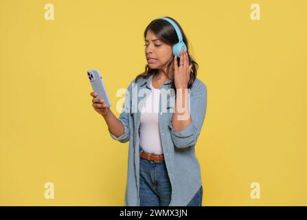 Unglückliche Indianerin, die auf ihr Handy schaut, Stereokopfhörer an den Ohren trägt, schlechte Nachrichten liest, in Jeans und Jeans gekleidet ist Stockfoto