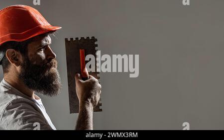 Renovierende Wand aus Gipskarton. Profil des bärtigen Baumeisters. Arbeiter mit Putzwerkzeugen. Renovierendes Hauskonzept. Baumeister-Mann im Schutzhelm. Werkzeug Stockfoto