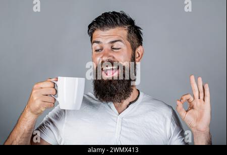 Glücklicher, lächelnder Mann, der eine Tasse leckeren frischen Kaffee hält und den Daumen nach oben zeigt. Der Mann hält eine Tasse Kaffee, Tee. Bärtiger Mann lächelt und zeigt Zeichen ok. Gut Stockfoto