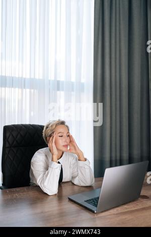 Vertikales Porträt einer müden Geschäftsfrau mittleren Alters, die ein Notebook benutzt und Probleme hat, aufgeregt über Fehler am Arbeitsplatz. Überarbeitete Unternehmerin Stockfoto