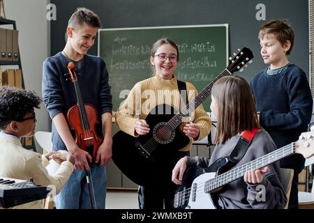 Gruppe fröhlicher Teenager-Jungen und -Mädchen mit Musikinstrumenten, die während der Probe im Klassenzimmer Pause machen Stockfoto