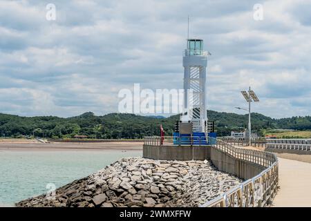 Weißer Leuchtturm am Ende des Piers unter bewölktem Himmel mit Strand im Hintergrund in Südkorea Stockfoto