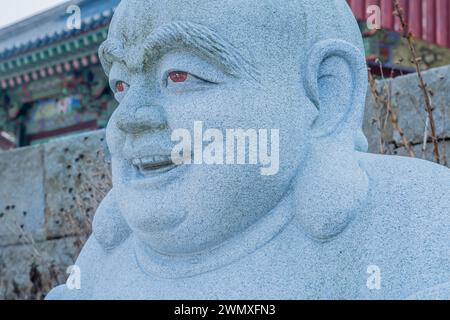 Nahaufnahme des Kopfes und des Gesichts des aus Stein geschnitzten sitzenden Buddha mit verschwommenem Tempelgebäude im Hintergrund in Pyeongtaek, Südkorea Stockfoto