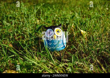 Ein Spielzeug einer überraschten blauen Eule mit mehrfarbigen Augen in Nahaufnahme steht mitten im grünen Gras im Licht der Abendsonne Stockfoto