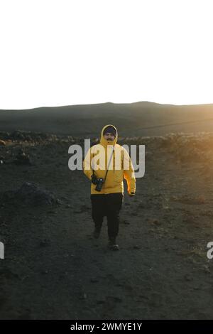 Ein Mann in einer leuchtend gelben Jacke mit einer Kamera wandert während des Sonnenuntergangs durch die vulkanische Landschaft Islands. Stockfoto