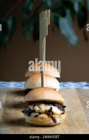 Drei Mini-Beef-Burger mit geschmolzenem Cheddar-Käse serviert auf einer rustikalen Holzplanke, die zur Präsentation aufgespießt werden. Stockfoto