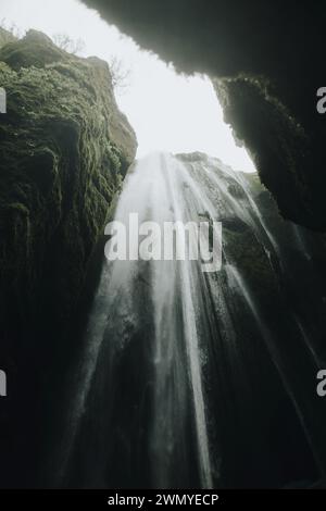 Von unten atemberaubende Aussicht auf einen mächtigen isländischen Wasserfall, der unter einem weichen, diffusen Sonnenlicht moosbedeckte Klippen herabstürzt. Stockfoto