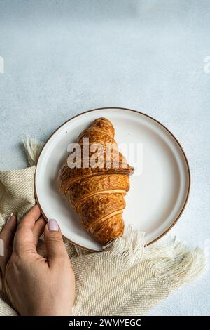 Blick von oben auf eine anonyme Person, die einen Teller mit einem frisch gebackenen Croissant auf einem strukturierten Tuch hält, was einen einfachen und gemütlichen Moment beim Frühstück festmacht Stockfoto