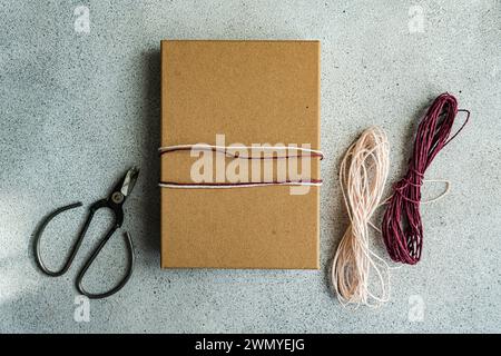 Ein einfaches, aber elegantes Geschenk in braunem Papier mit rosa und weißem Garn, neben einer Schere und zusätzlichem Garn auf strukturiertem grauen Hintergrund Stockfoto