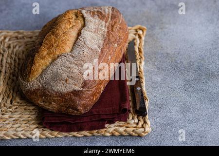 Ein knuspriger Brotlaib mit selbstgemachtem Roggensauerteig, auf einer gewebten Matte mit einer dunkelroten Serviette und einem Brotmesser Stockfoto